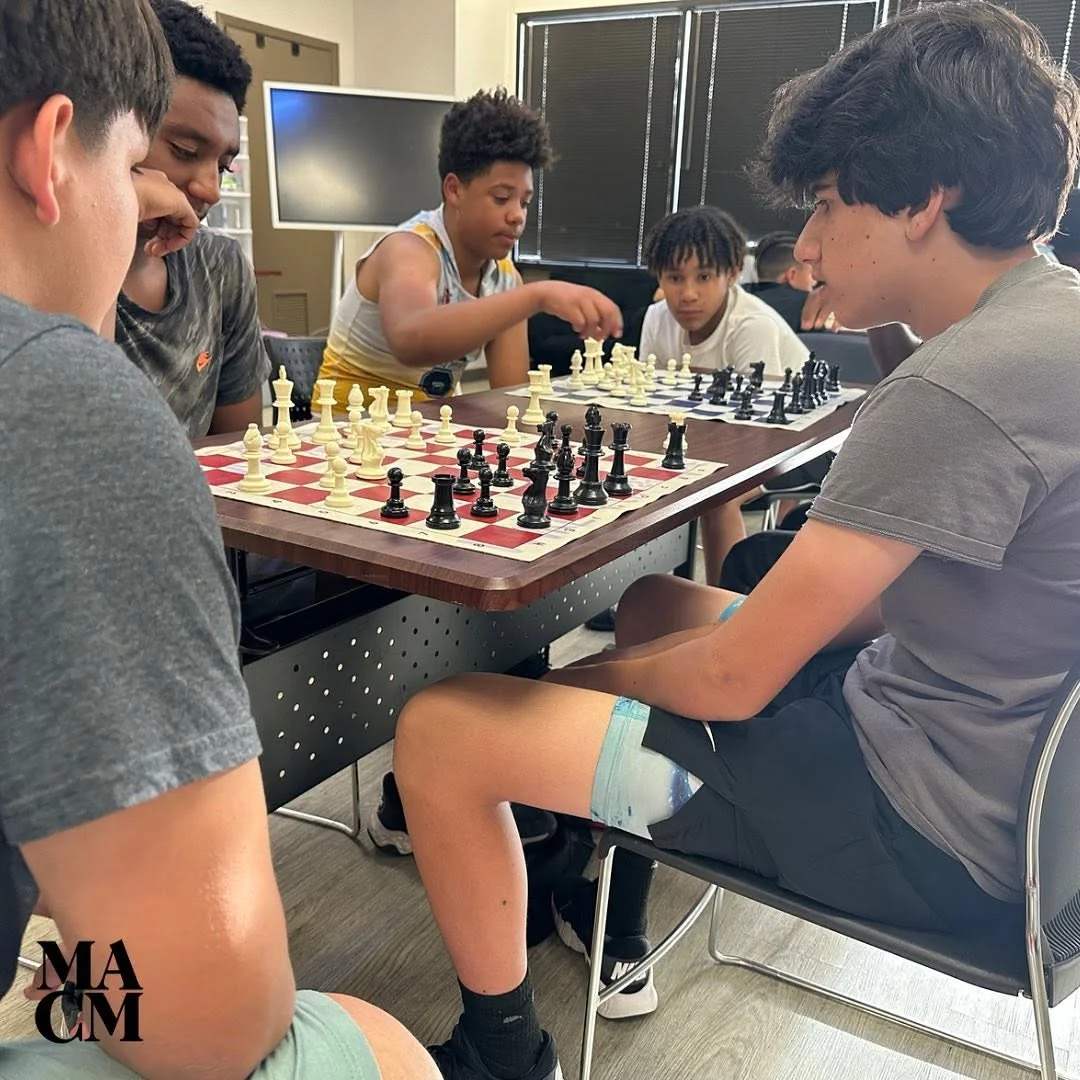 A group of youth gathered around chess boards in a MACM learning space